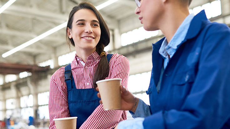 Positive young female construction workers drinking coffee from disposable cups and talking while resting during break