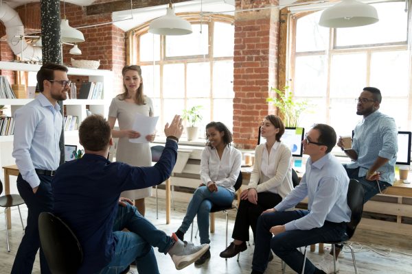 Business people in a casual modern office, gathered together, one male has his hand raised to ask the mentor a question