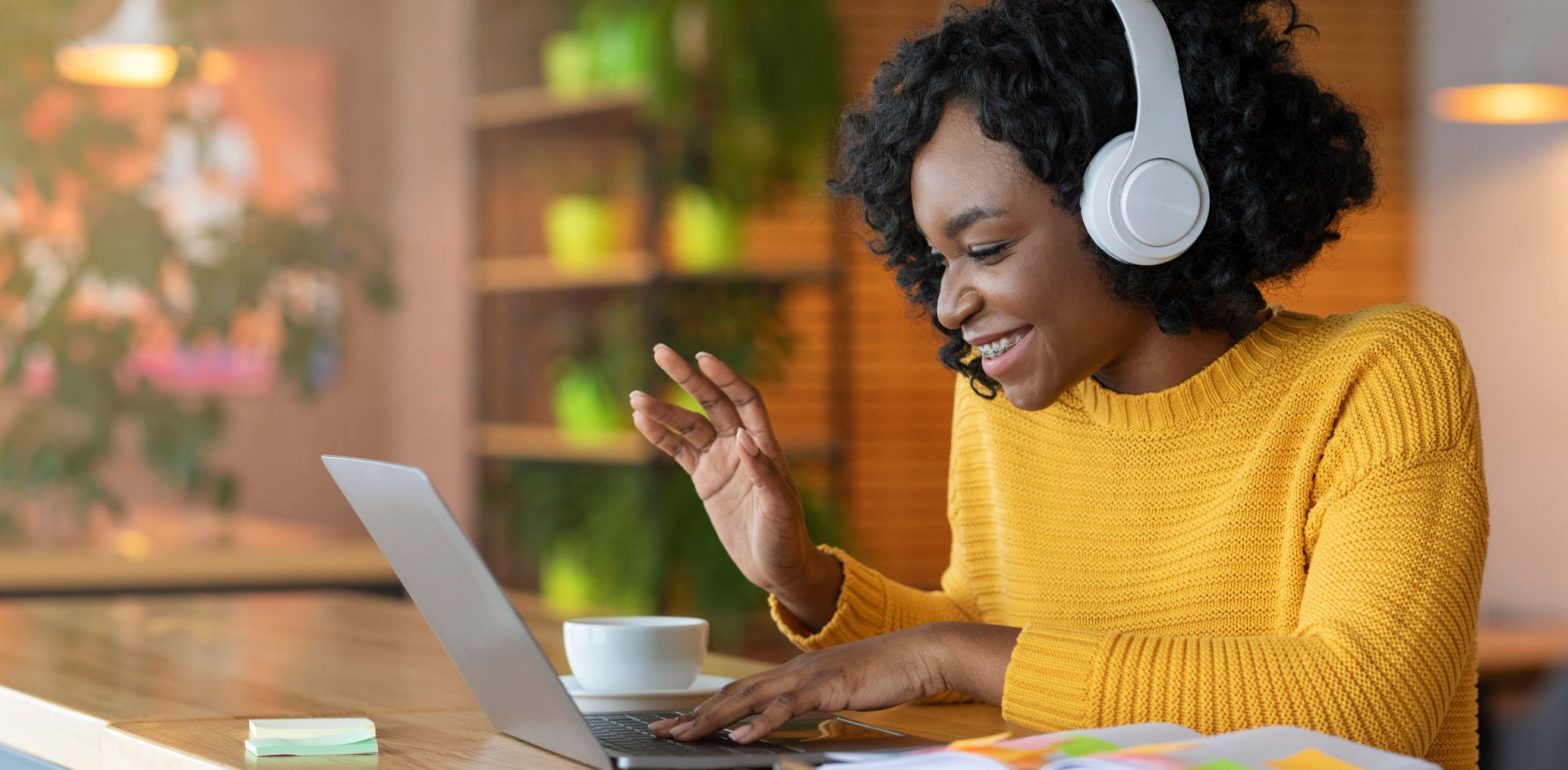 A woman wearing a yellow jumper and white headphones is smiling and working on her laptop.