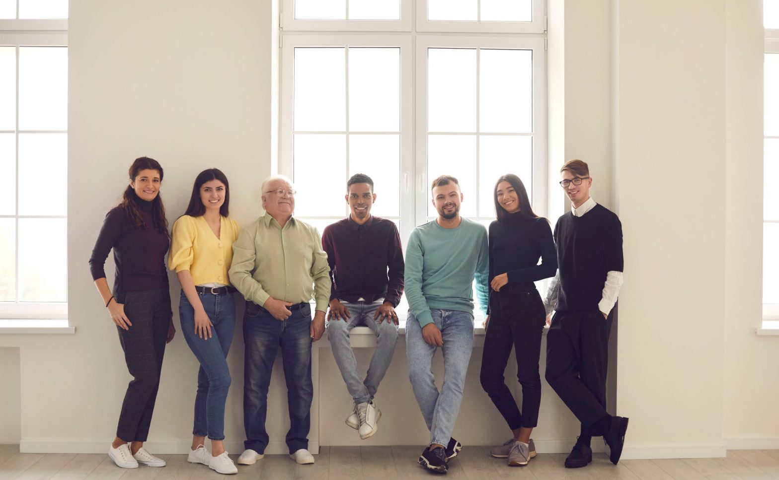 Group of man and women of mixed ages posing together in front of windows.