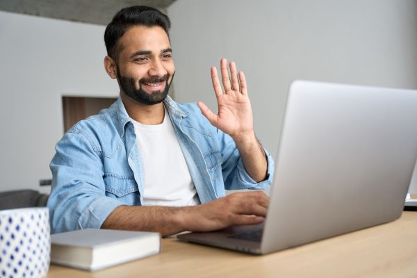 A man is smiling and waving at the inbuilt camera in his laptop