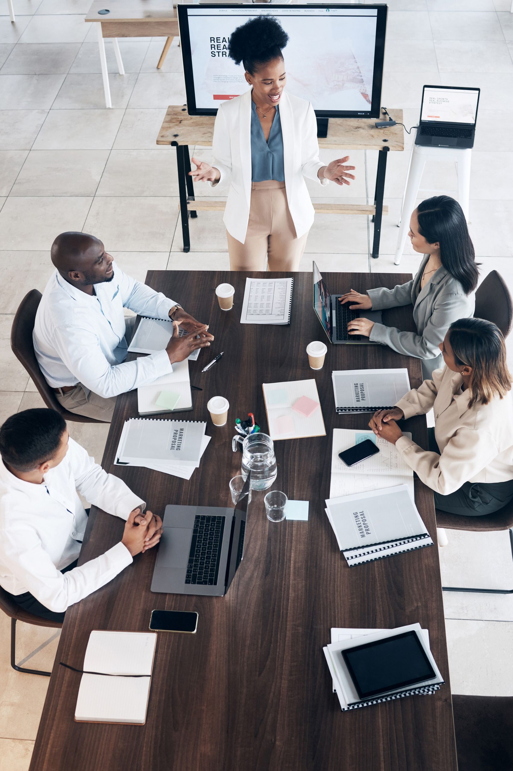 Business men and women sitting at a table with laptops and notebooks with a mentor giving a presentation