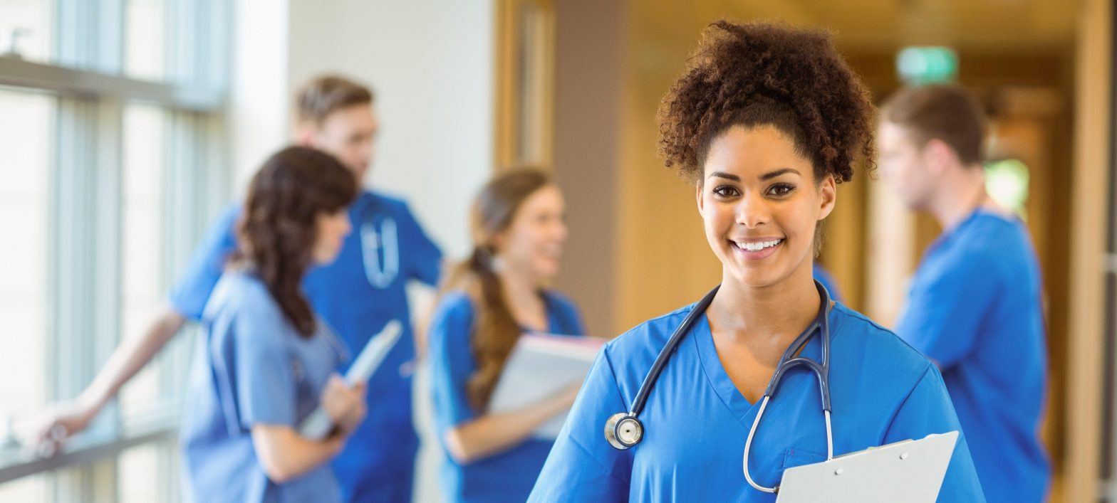 Female medical professional wearing blue scrubs smiling at the camera while her colleagues are having a conversation behind her.