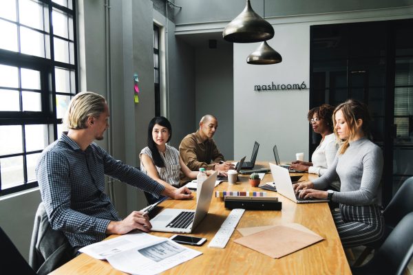 Business people sit around a large desk working on their laptops and pausing for discussion.