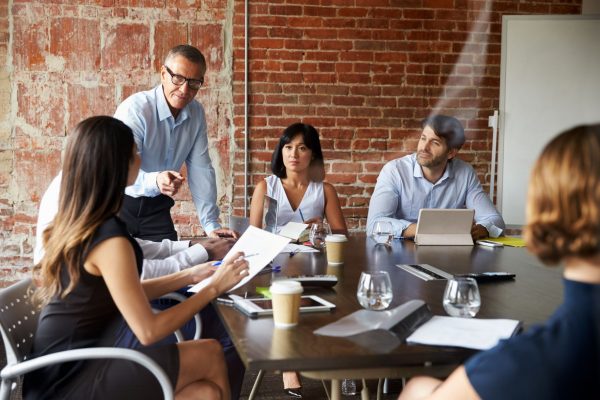 A business mentor is leading a discussion around a table and pointing at a female colleague who is holding notes and wishes to contribute.