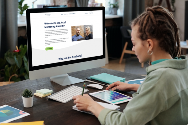 The Art of Mentoring website is displayed on a desktop computer and a woman is making notes