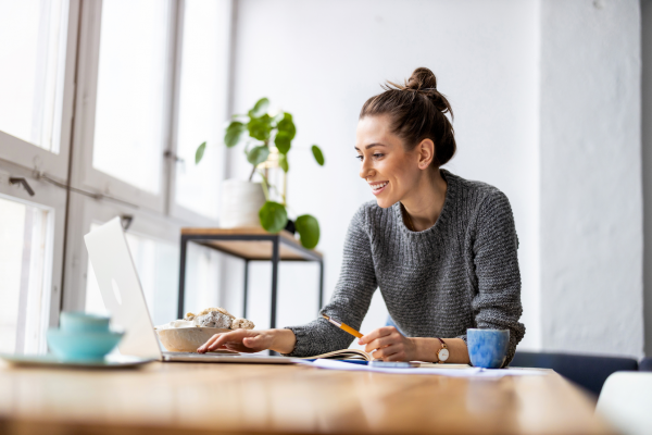 Smiling female wearing a grey jumper and with her hair in a bun is working on a laptop.