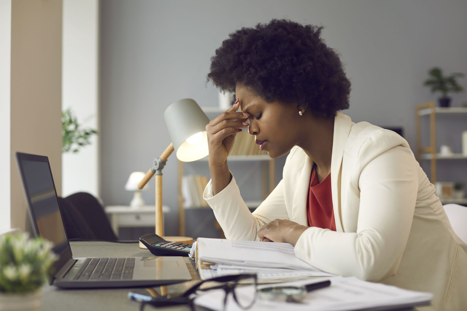 A frustrated businesswoman holds her forehead as she is surrounded by paperwork and her laptop.