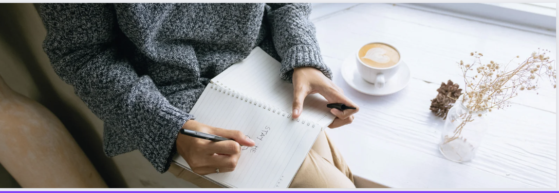 A woman wearing a grey jumper is writing in a notebook which is balanced on her knee. She has a coffee and a plant beside her.