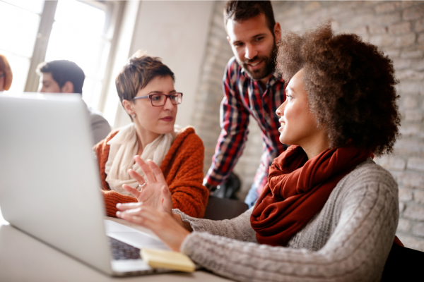 Colleagues are in discussion and a mentor stands over them participating in the conversation.