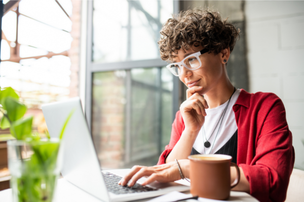 Young female with short curly hair and white glasses is working on her laptop with a coffee beside her