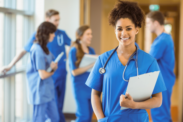 Female medical professional wearing blue scrubs smiling at the camera while her colleagues are having a conversation behind her.