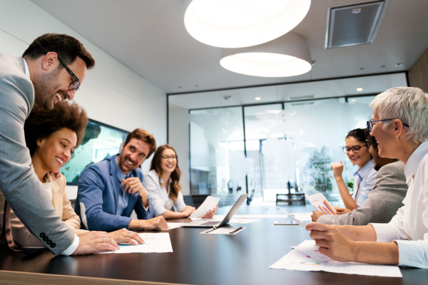 A mentor is leading a group session around a conference table. Business men and women sit around the table smiling and listening to the mentor.